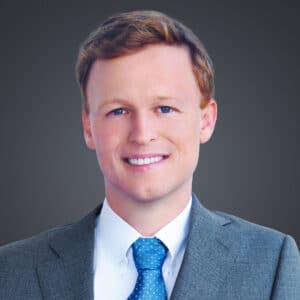 A professional headshot of Dr. Crist in a gray suit. He is wearing a white collared shirt and a blue tie with white dots, and is looking directly at the camera. The background is a solid dark gray.