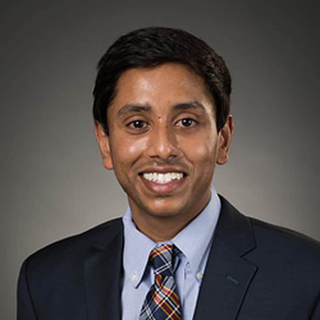 Professional headshot of a smiling man with dark hair wearing a dark suit, light blue shirt, and plaid tie, against a gray studio background
