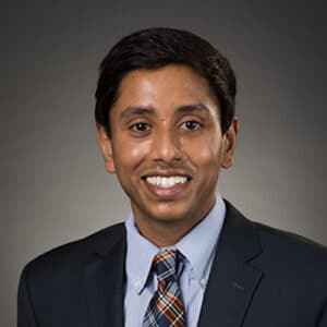 Professional headshot of a smiling man with dark hair wearing a dark suit, light blue shirt, and plaid tie, against a gray studio background